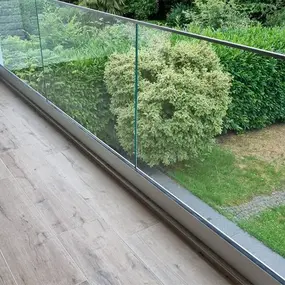 Balkon mit modernem Glasgeländer und Holzoptik-Fliesen, Blick auf grünen Garten.