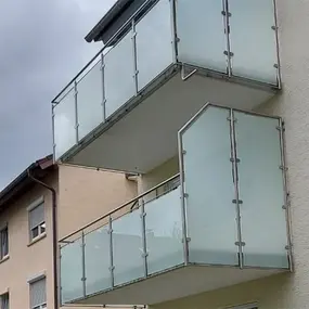 Zwei moderne Balkone mit Milchglas-Geländern an einer hellen Hausfassade bei bewölktem Himmel.