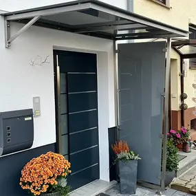 Moderne Haustür mit Vordach aus Glas, Briefkasten und Blumen vor einem Wohnhaus.