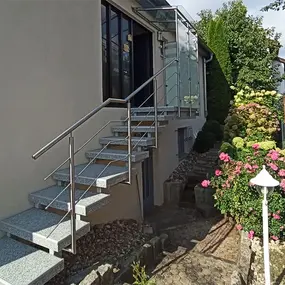 Außentreppe aus Granit mit Edelstahlgeländer an einem Haus, umgeben von Garten und Blumen.