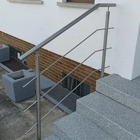 Modernes Edelstahlgeländer an einer Außentreppe mit Granitstufen vor einem Wohnhaus.