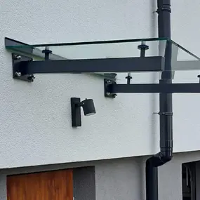 Modernes Vordach aus Glas und Metall an einer Hauswand mit Regenrinne und Außenleuchte.