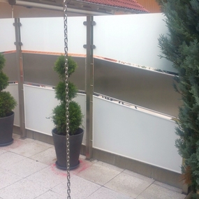 Moderner Sichtschutz aus Glas und Metall auf Balkon mit Pflanzen und hellen Bodenfliesen.