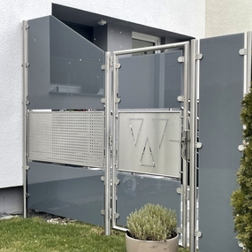 Moderner Sichtschutz aus Glas und Edelstahl mit Tür und W-Logo vor einem Haus.