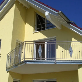 Modernes Einfamilienhaus mit Balkon, gelber Fassade und roten Dachziegeln bei Sonnenschein.