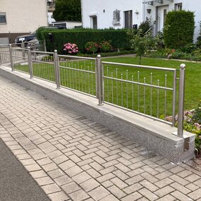Moderner Edelstahlzaun mit Steinmauer vor gepflegtem Vorgarten und Einfamilienhaus.